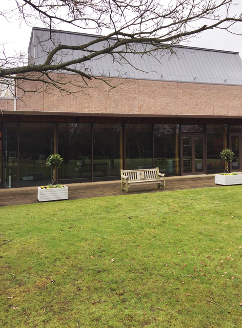 Color photo of the entrance to the concert hall. It is a modern structure, with a glass wall for the entrance lobby area. A bench and two planter boxes sit on the brick front walkway, with a lawn area in the foreground.